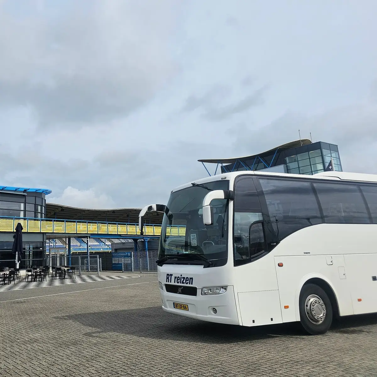 Touringcar van RT Reizen staat voor de tribune van een circuit, met de wedstrijdtoren erachter.