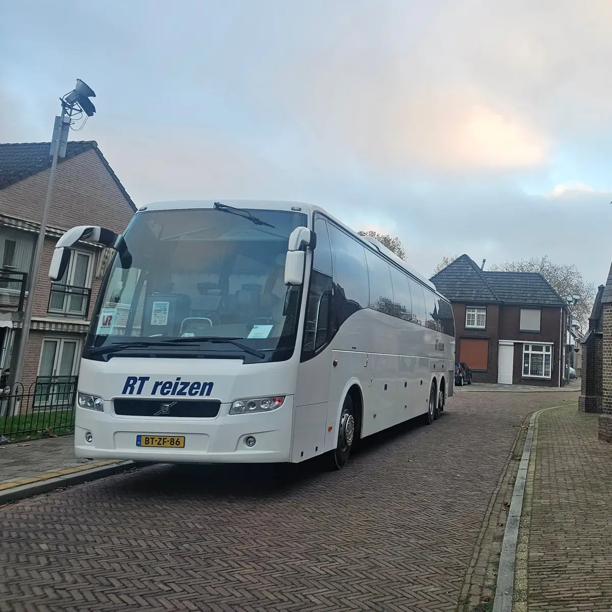 Touringcar van RT Reizen staat in een dorpsstraat op klinkers tussen de huizen, met de hele bus in beeld.