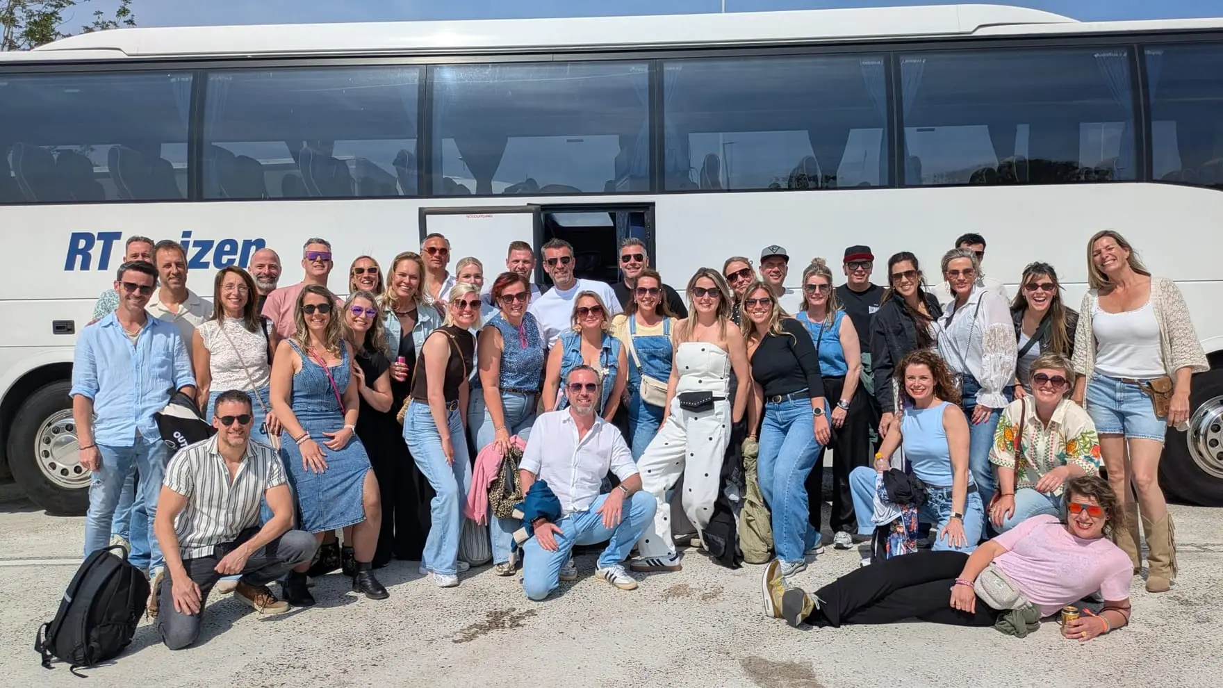 Een groep collega's staat lachend voor de touringcar van RT Reizen, op een zonnige dag